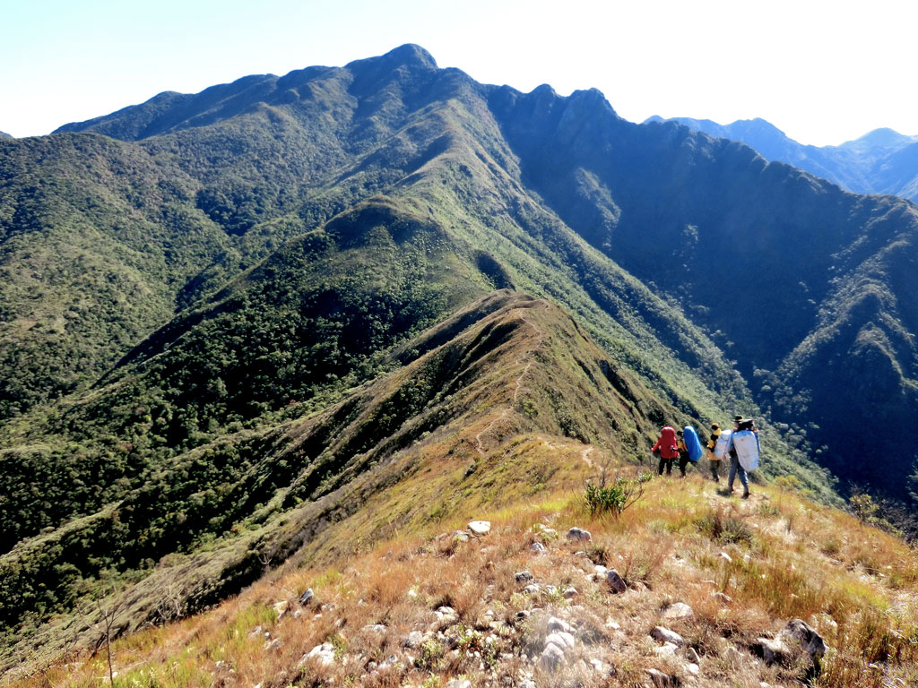 Andarilhos caminham pela trilha da Serra Fina rumo ao Alto do Capim Amarelo (2.570 m), na Serra da Mantiqueira. A próxima parada nesta travessia é a Pedra da Mina (atrás), a quarta montanha mais alta do Brasil, com 2.798 m.